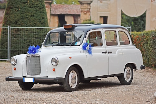 A classic white car, resembling a vintage taxi, is adorned with blue ribbon decorations on its front and side mirrors. It is parked on a gravel surface with greenery and an old building in the background.