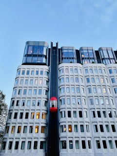 A modern building with a unique facade featuring a grid of windows and a prominent red elevator moving vertically along the exterior. The upper levels are constructed with sleek, reflective glass panels. The sky is clear and blue, indicating a pleasant day.