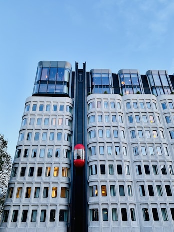 A modern building with a unique facade featuring a grid of windows and a prominent red elevator moving vertically along the exterior. The upper levels are constructed with sleek, reflective glass panels. The sky is clear and blue, indicating a pleasant day.
