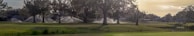 A lush landscape featuring a well-maintained golf course with large trees in the foreground. Sprinklers are actively watering the grass, creating arcs of water. In the distance, there's a small pond and golf carts suggesting leisure and recreation activities.
