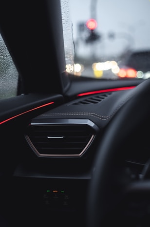 A car dashboard is seen with an air vent and red ambient lighting accentuating the interior. Raindrops are visible on the window, and blurred traffic lights can be seen in the background, adding a soft glow to the scene.