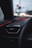 A car dashboard is seen with an air vent and red ambient lighting accentuating the interior. Raindrops are visible on the window, and blurred traffic lights can be seen in the background, adding a soft glow to the scene.