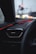 A car dashboard is seen with an air vent and red ambient lighting accentuating the interior. Raindrops are visible on the window, and blurred traffic lights can be seen in the background, adding a soft glow to the scene.