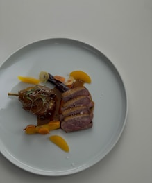 A gourmet dish presented on a white plate featuring slices of cooked duck breast with a crispy skin, garnished with herbs. Accompanied by a caramelized piece of meat with bone, likely lamb or duck leg, and various sliced vegetables such as orange segments and carrots.