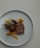 A gourmet dish presented on a white plate featuring slices of cooked duck breast with a crispy skin, garnished with herbs. Accompanied by a caramelized piece of meat with bone, likely lamb or duck leg, and various sliced vegetables such as orange segments and carrots.