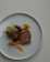 A gourmet dish presented on a white plate featuring slices of cooked duck breast with a crispy skin, garnished with herbs. Accompanied by a caramelized piece of meat with bone, likely lamb or duck leg, and various sliced vegetables such as orange segments and carrots.