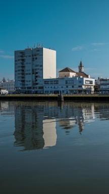 A cityscape featuring a large white apartment building and a church with a steeple beside a calm body of water. The buildings are clearly reflected in the water, and the sky is a clear blue, indicating a sunny day.