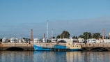 A small blue and yellow boat is docked beside a concrete pier. The pier has several arches beneath it, and a row of large white trucks is parked along the top. Behind the trucks, various trees and buildings are visible, including some tall brick chimneys. The sky above is clear with a few clouds, and the water in the foreground is calm, reflecting the boat and pier.
