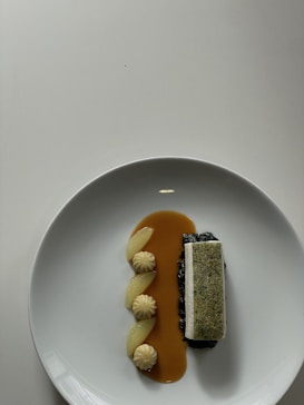 A neatly plated dish on a white plate. The dish has a rectangular piece topped with a crust on a bed of dark green vegetables accompanied by a smooth brown sauce. On the side, there are three dollops of creamy puree and three segments of a yellow citrus fruit.