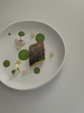 A minimalist plate of food featuring a piece of fish with crispy skin, served with creamy elements and small green circular garnishes. There are also cubes and a small vibrant flower, all presented on a plain white plate.