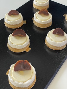 Several round pastries are arranged on a black serving tray. Each pastry has a light cream filling topped with a decorative brown chocolate piece. The surface of the pastries is smooth and creamy, with a slightly darker crust at the bottom.