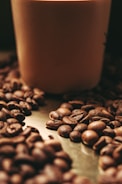 A collection of coffee beans is scattered on a metallic surface, creating a rustic and cozy atmosphere. A soft focus effect adds a warm ambiance to the scene, with a large cylindrical object, possibly a candle or container, in the background.