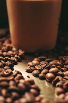 A collection of coffee beans is scattered on a metallic surface, creating a rustic and cozy atmosphere. A soft focus effect adds a warm ambiance to the scene, with a large cylindrical object, possibly a candle or container, in the background.