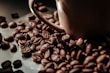 A close-up view of roasted coffee beans scattered around a brown ceramic coffee cup. The beans have a rich, dark brown color and a glossy finish, suggesting freshness and quality.