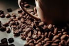 A close-up view of roasted coffee beans scattered around a brown ceramic coffee cup. The beans have a rich, dark brown color and a glossy finish, suggesting freshness and quality.