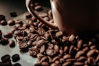 A close-up view of roasted coffee beans scattered around a brown ceramic coffee cup. The beans have a rich, dark brown color and a glossy finish, suggesting freshness and quality.
