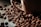 A close-up view of roasted coffee beans scattered around a brown ceramic coffee cup. The beans have a rich, dark brown color and a glossy finish, suggesting freshness and quality.