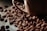 A close-up view of roasted coffee beans scattered around a brown ceramic coffee cup. The beans have a rich, dark brown color and a glossy finish, suggesting freshness and quality.