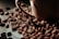 A close-up view of roasted coffee beans scattered around a brown ceramic coffee cup. The beans have a rich, dark brown color and a glossy finish, suggesting freshness and quality.