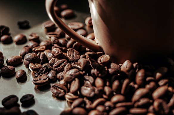 A close-up view of roasted coffee beans scattered around a brown ceramic coffee cup. The beans have a rich, dark brown color and a glossy finish, suggesting freshness and quality.