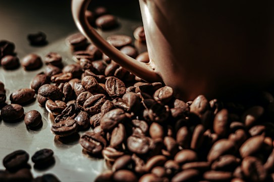 A close-up view of roasted coffee beans scattered around a brown ceramic coffee cup. The beans have a rich, dark brown color and a glossy finish, suggesting freshness and quality.