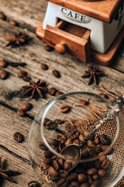 A rustic scene featuring a vintage coffee grinder labeled 'Café', surrounded by scattered coffee beans and star anise on a wooden surface. A glass cup partially filled with coffee beans is placed in the foreground, accompanied by a small metal spoon resting inside.