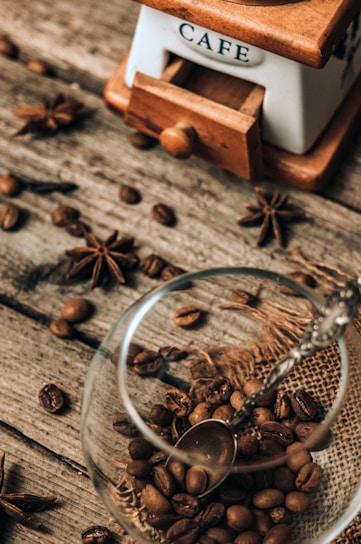 A rustic scene featuring a vintage coffee grinder labeled 'Café', surrounded by scattered coffee beans and star anise on a wooden surface. A glass cup partially filled with coffee beans is placed in the foreground, accompanied by a small metal spoon resting inside.
