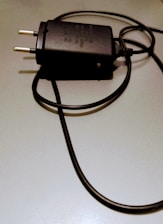 A black electrical plug with a coiled cord is placed on a light-colored surface. The plug has two metal prongs and features some text or labeling on its body.