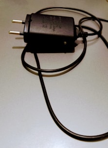 A black electrical plug with a coiled cord is placed on a light-colored surface. The plug has two metal prongs and features some text or labeling on its body.