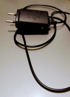 A black electrical plug with a coiled cord is placed on a light-colored surface. The plug has two metal prongs and features some text or labeling on its body.
