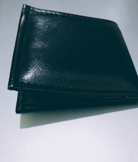 A closed black leather wallet is placed on a flat, light-colored surface. The wallet appears to have a smooth texture with some shine reflecting off its surface.