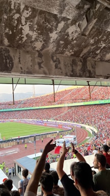 A crowded football stadium filled with cheering fans. People are waving flags and raising their hands, showing enthusiasm and excitement. The stadium's architecture is visible with a worn concrete ceiling. On the field, a green football pitch is visible, surrounded by a running track. Security personnel are present along the perimeter.