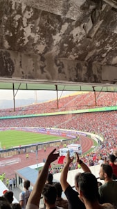 A crowded football stadium filled with cheering fans. People are waving flags and raising their hands, showing enthusiasm and excitement. The stadium's architecture is visible with a worn concrete ceiling. On the field, a green football pitch is visible, surrounded by a running track. Security personnel are present along the perimeter.