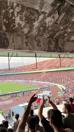 A crowded football stadium filled with cheering fans. People are waving flags and raising their hands, showing enthusiasm and excitement. The stadium's architecture is visible with a worn concrete ceiling. On the field, a green football pitch is visible, surrounded by a running track. Security personnel are present along the perimeter.