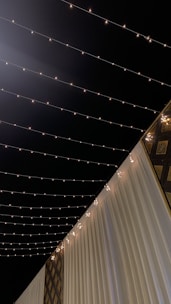 A nighttime scene with string lights hanging overhead, creating a warm and inviting atmosphere. The lights are arranged in parallel lines across the dark sky. Below, there are elegant white drapes and decorative gold patterns adding a touch of luxury.
