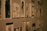 A wall covered with multiple black and white sketches or drawings arranged in a grid-like pattern. The illustrations vary in style and subject matter, and some appear to depict human figures and abstract designs. The lighting is dim, contributing to a moody atmosphere.