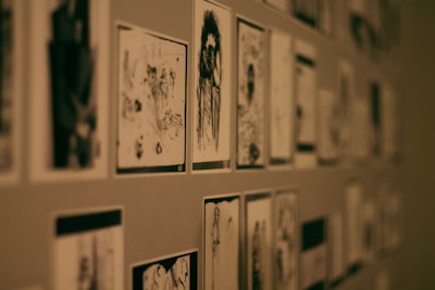 A wall covered with multiple black and white sketches or drawings arranged in a grid-like pattern. The illustrations vary in style and subject matter, and some appear to depict human figures and abstract designs. The lighting is dim, contributing to a moody atmosphere.
