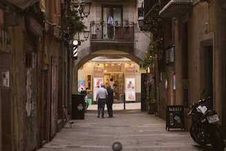 A narrow, cobblestone street flanked by tall, old buildings leads to a warmly lit shop. The shop showcases vibrant artwork in its windows. Two people walk down the street towards the shop entrance beneath an arched passageway. Potted plants and a few small lights hang from the buildings. A motorcycle is parked on the side, and there's a barber pole indicating a nearby barber shop.