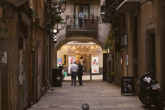 A narrow, cobblestone street flanked by tall, old buildings leads to a warmly lit shop. The shop showcases vibrant artwork in its windows. Two people walk down the street towards the shop entrance beneath an arched passageway. Potted plants and a few small lights hang from the buildings. A motorcycle is parked on the side, and there's a barber pole indicating a nearby barber shop.