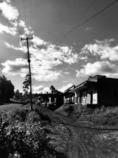 A rural road flanked by foliage leads to a series of small structures or houses. Overhead, electricity lines stretch across the sky which is filled with scattered clouds, creating a tranquil setting. The road and vegetation suggest a remote area potentially in a village or countryside.