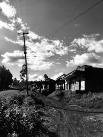A rural road flanked by foliage leads to a series of small structures or houses. Overhead, electricity lines stretch across the sky which is filled with scattered clouds, creating a tranquil setting. The road and vegetation suggest a remote area potentially in a village or countryside.