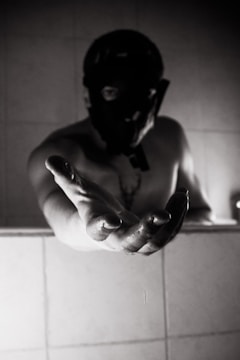 A person wearing a mask extends their hand towards the viewer. The lighting creates strong shadows, adding to the mystery. The figure is partially obscured by darkness, and the background consists of a tiled wall.