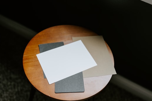 A small wooden round table holds three sheets of paper. The sheets are different colors, including white, gray, and beige. The background is dark, adding contrast to the table and papers.