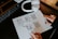 A person is drawing a flowchart on a dotted notepad. The flowchart includes boxes and arrows, possibly representing a design or planning process. A pair of white headphones rests nearby on the dark desk surface. The person’s nails are painted dark, and they are wearing rings.