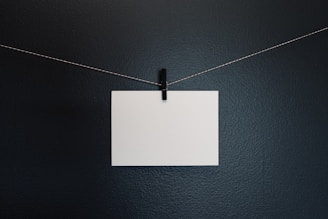 A blank white piece of paper is attached to a thin, string-like wire with a small black clothespin. The background is a solid, dark gray surface.