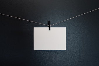 A blank white piece of paper is attached to a thin, string-like wire with a small black clothespin. The background is a solid, dark gray surface.