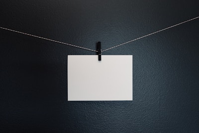 A blank white piece of paper is attached to a thin, string-like wire with a small black clothespin. The background is a solid, dark gray surface.