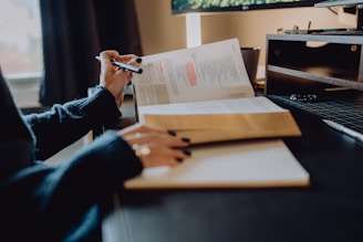 A person is sitting at a desk and reading a book with highlighted text. There is a second book open nearby and a notepad in the foreground. The setting appears to be a cozy, well-organized workspace with a computer monitor and shelving unit in the background.