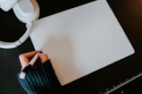 A person wearing a dark sweater is holding a pen and writing on a blank sheet of white paper. Nearby, there is a pair of white headphones and a ruler. The surface is dark, providing contrast with the bright paper and objects.