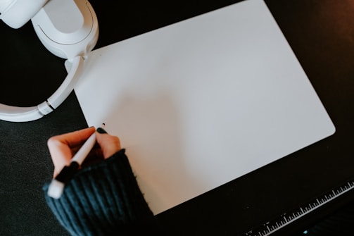 A person wearing a dark sweater is holding a pen and writing on a blank sheet of white paper. Nearby, there is a pair of white headphones and a ruler. The surface is dark, providing contrast with the bright paper and objects.